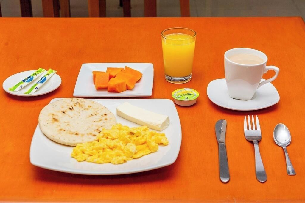 Desayuno con huevos revueltos, una porción de pan plano, una loncha de queso, jugo de naranja en un vaso, una taza de café y un recipiente pequeño de mantequilla.