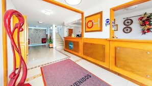 A hotel reception area with a reception desk, a red abstract sculpture, a clock, and a 'WELCOME' mat.