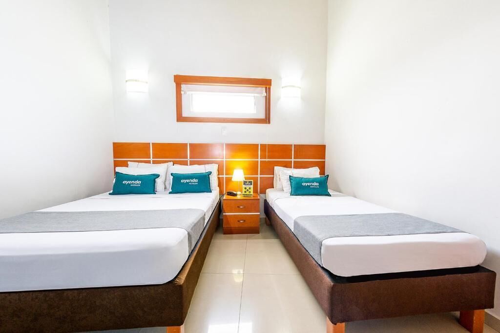 A hotel room with two beds, a nightstand, a wall-mounted lamp, and a window above the beds.