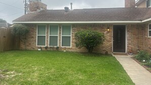 A single-story brick house with a front yard, a wooden fence, and a small tree.