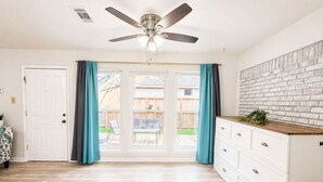 A room with a ceiling fan, a white door, a bench, a dining table, and a kitchen area with a brick wall.