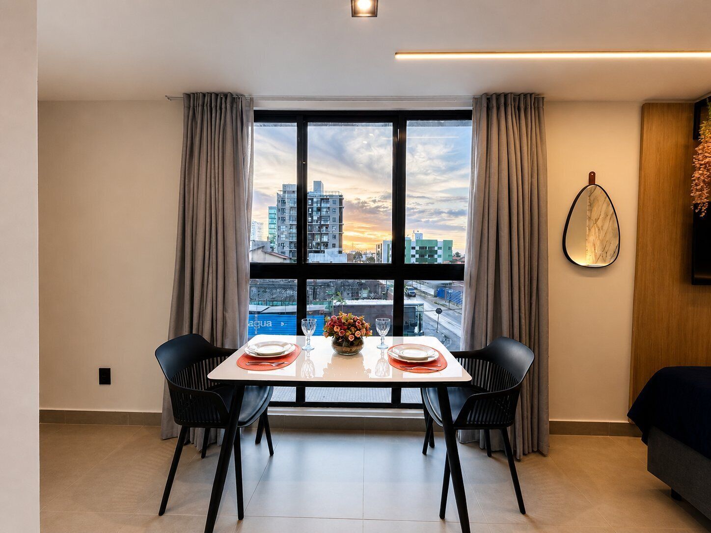 A dining area with a table set for two, a city view through large windows, and a modern interior design.