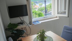 Une pièce avec une table en bois, une plante en pot, un téléviseur fixé au mur et une fenêtre donnant sur un balcon et de la verdure.