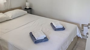 Un lit bien fait, avec des draps blancs et deux serviettes pliées, une lampe de chevet et une porte dotée d’une poignée moderne.