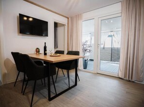 Ein moderner Essbereich mit Holztisch und schwarzen Stühlen, einem an der Wand befestigten Flachbildfernseher und einer Glastür, die auf einen Außenbalkon führt.
