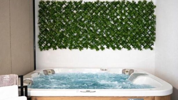Um quarto de spa com uma banheira de madeira, uma parede verde com padrão de folhas e uma toalha branca no chão.