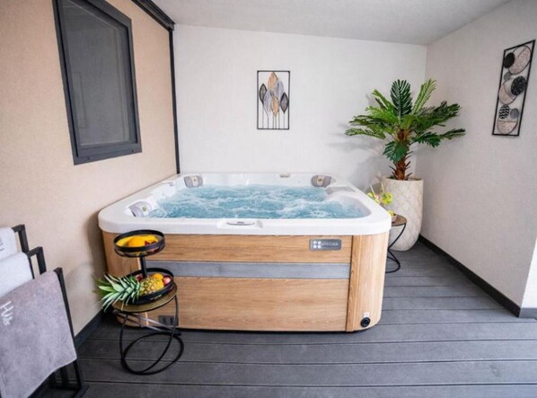 A hot tub in a room with a wooden floor, a small table with fruits, a potted plant, and a framed picture on the wall.
