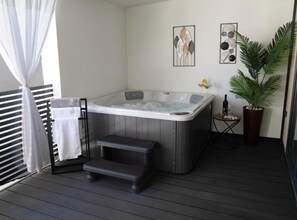 Ein Whirlpool auf einer Holzterrasse mit Handtuchhalter, einem kleinen Tisch mit Flasche und Glas sowie einem Blumentopf.
