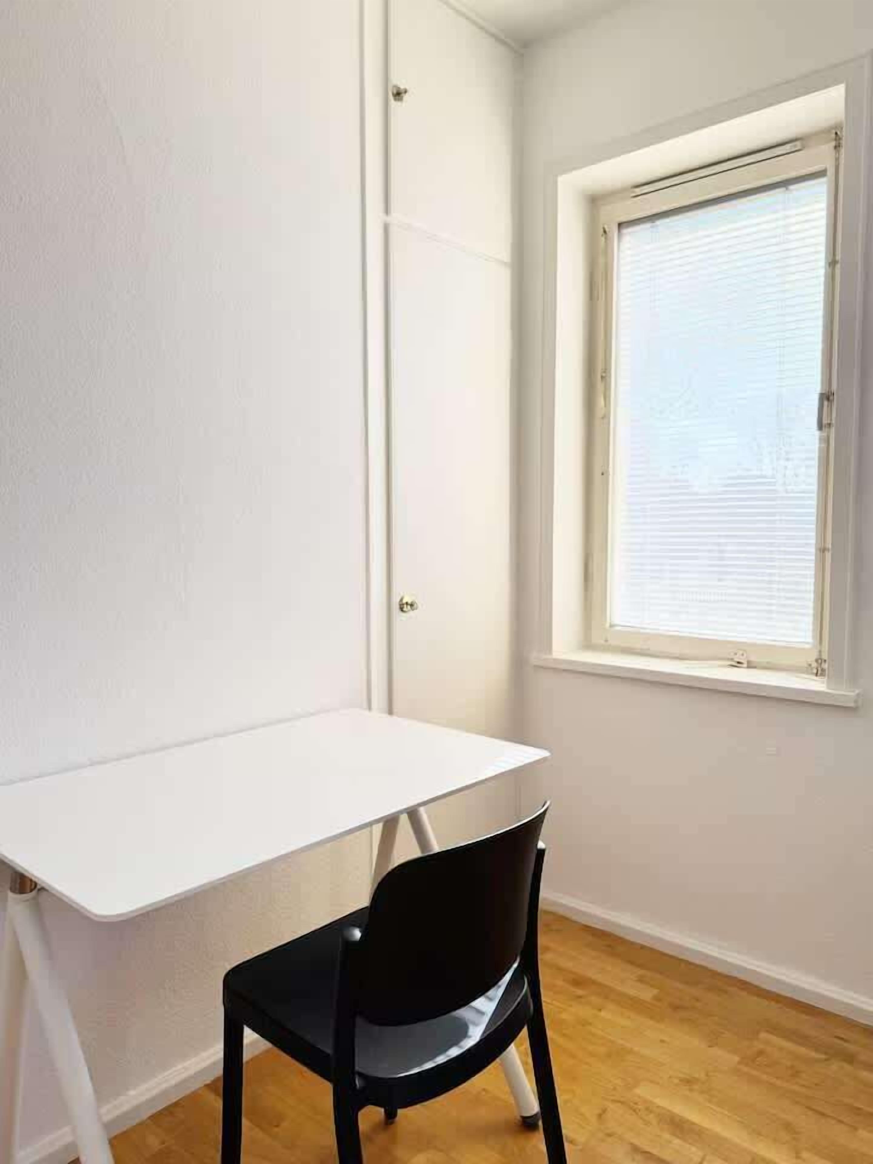 A room with a white desk, a black chair, a window with blinds, and wooden flooring.