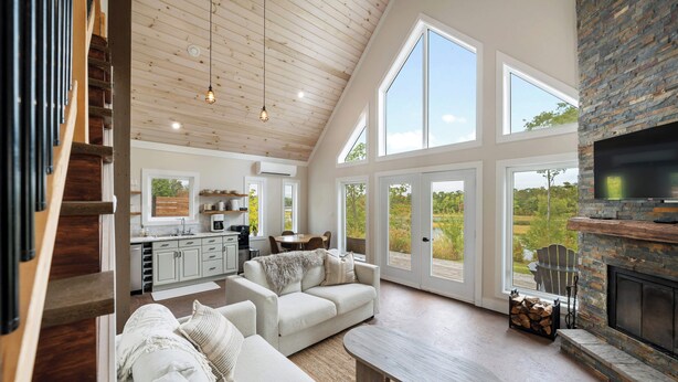 Un amplio salón con techo alto, ventanas grandes, chimenea y una cocina al fondo.