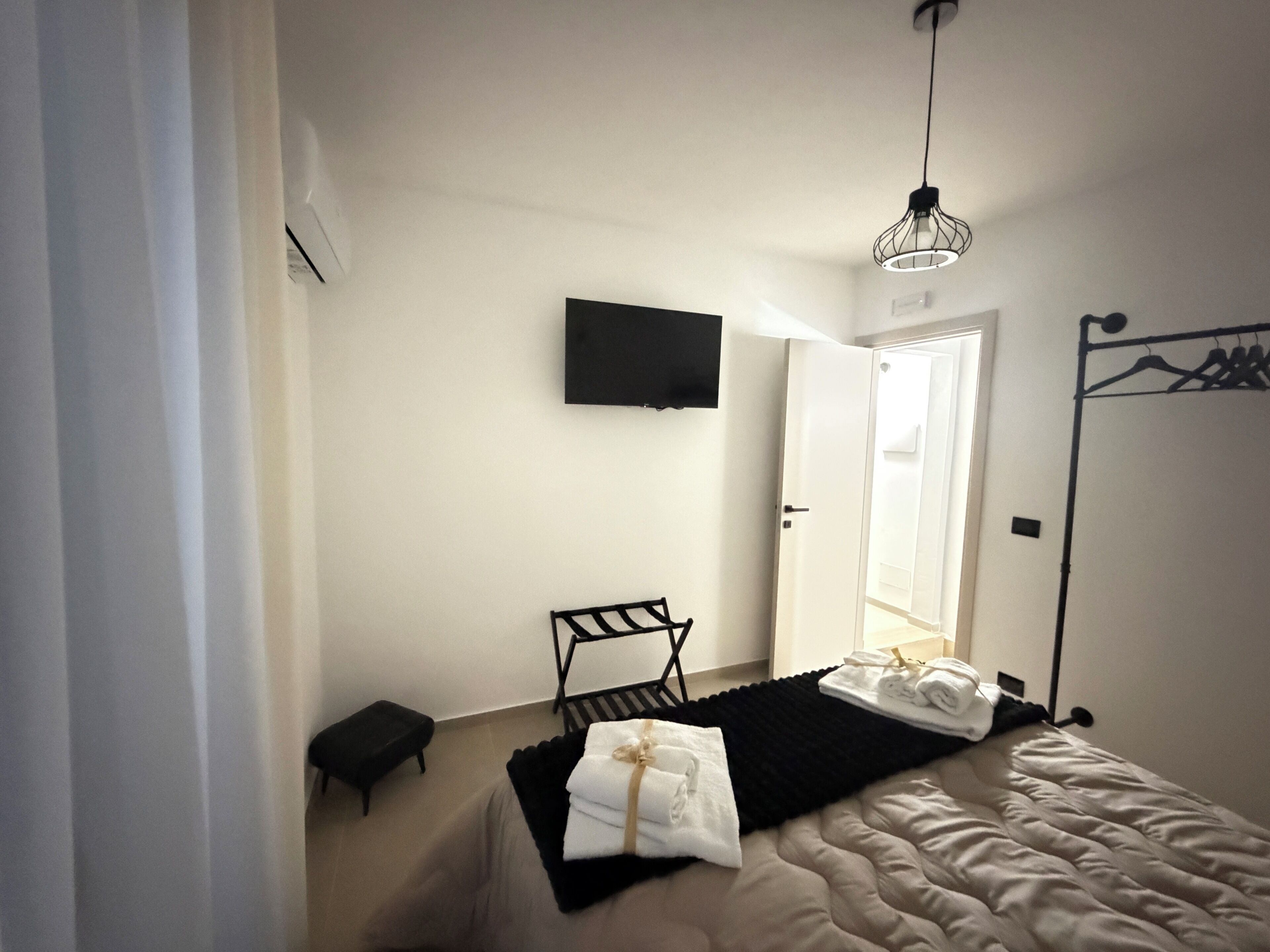 A bedroom with a bed, a chair, a small table, a TV mounted on the wall, and a door leading to another room.