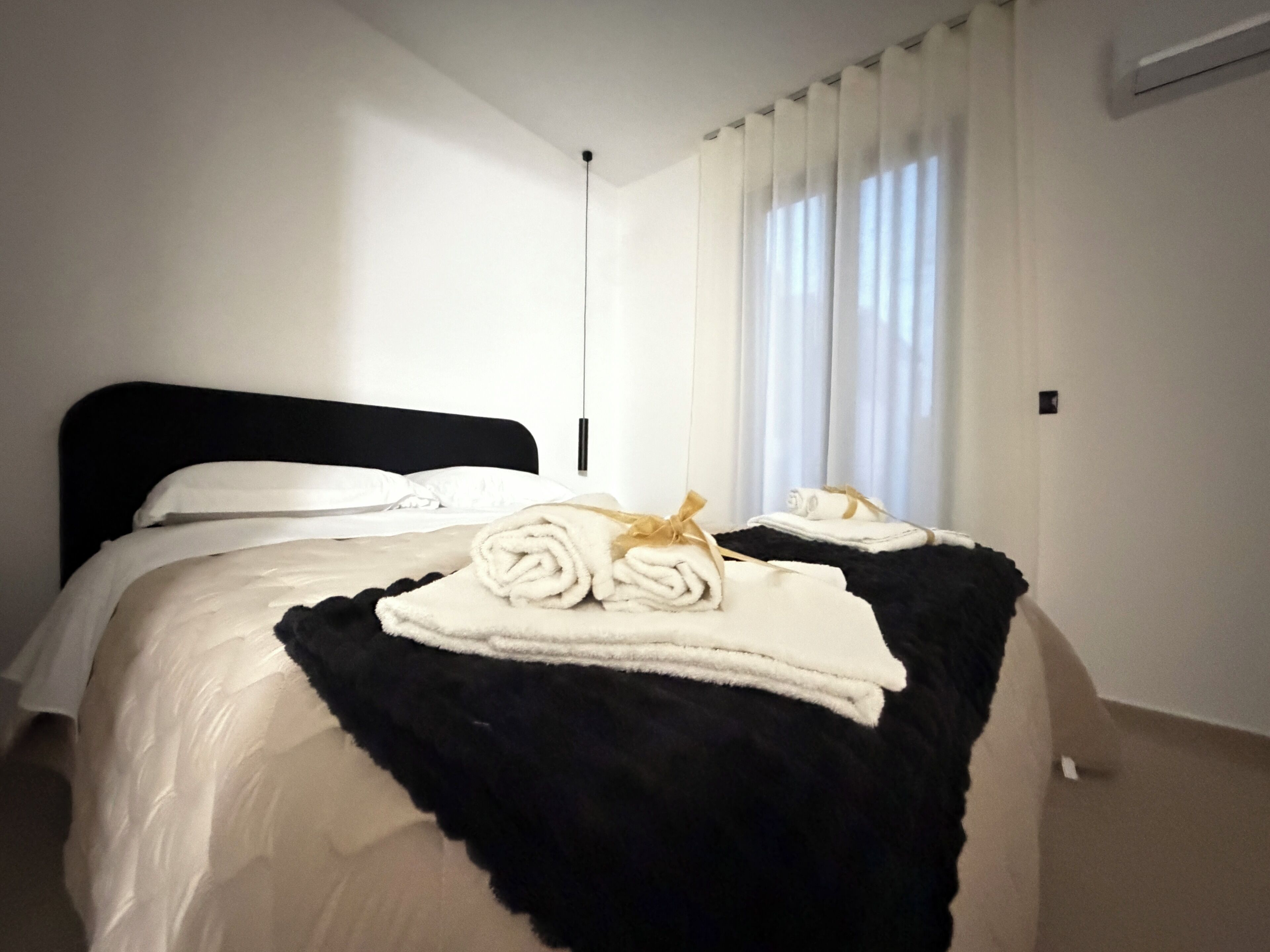 Camera d'albergo con un letto, una testiera nera, biancheria da letto bianca, una coperta a quadri bianconeri e una finestra con tende bianche.