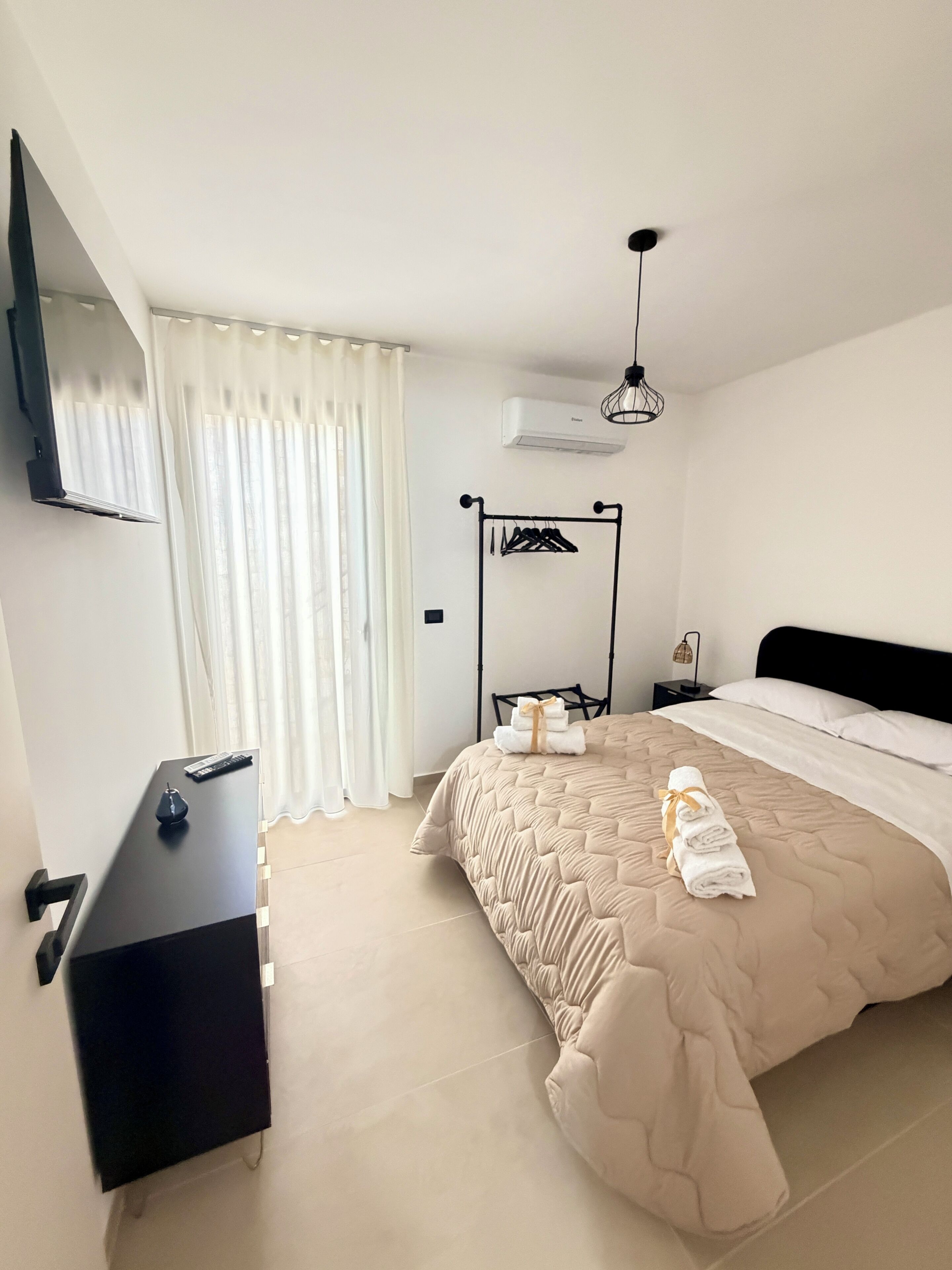 A bedroom with a bed, a desk, a TV, a wardrobe, a hanging light fixture, and a window with curtains.