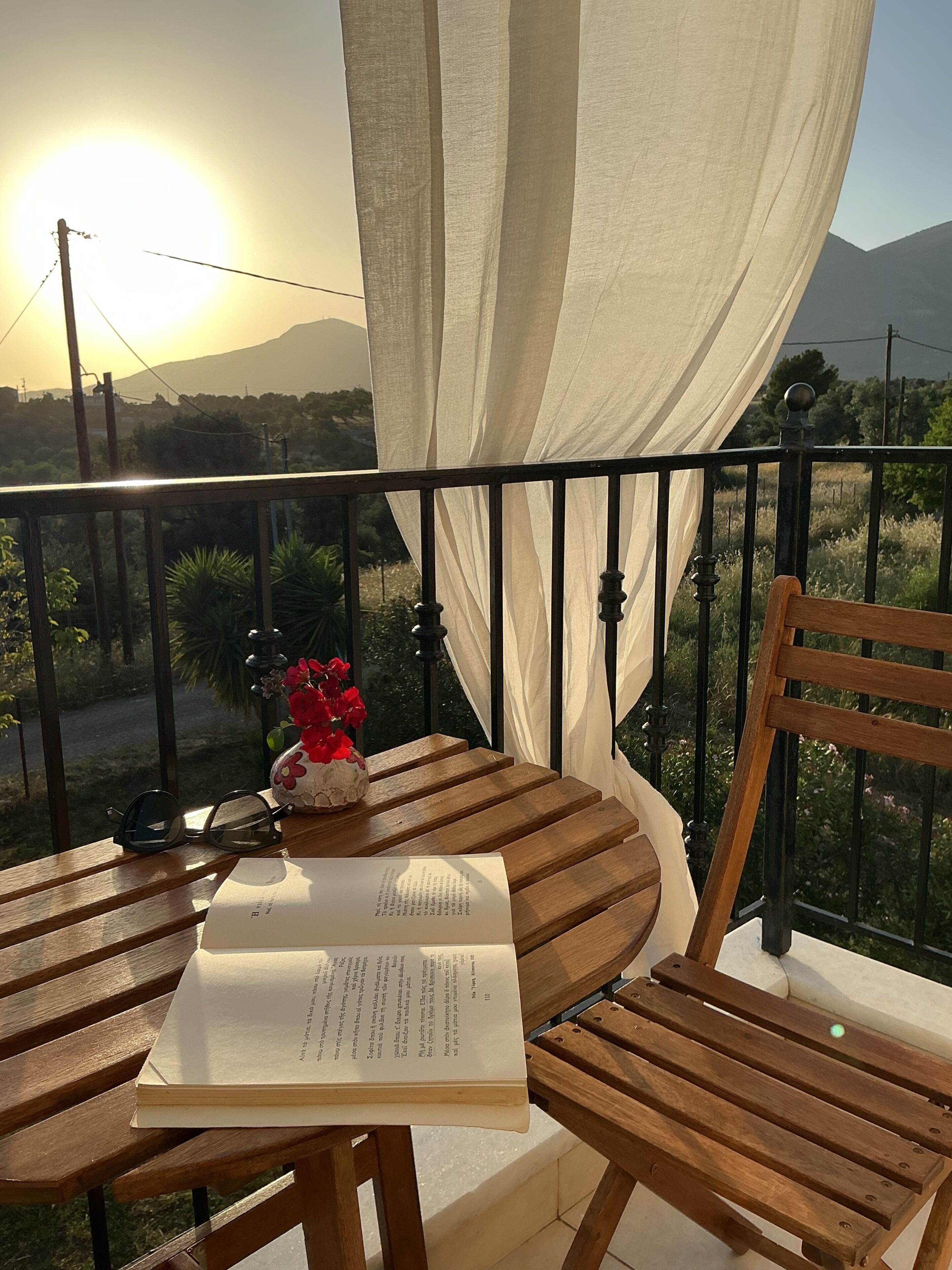 Una mesa de madera con un libro y un jarrón de flores rojas en un balcón con vista al sol y las montañas.