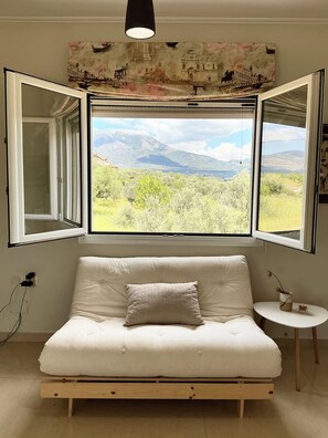 Ein Zimmer mit einem weißen Sofa, einem kleinen Beistelltisch und einem großen Fenster mit Blick auf Berge und Grünflächen.