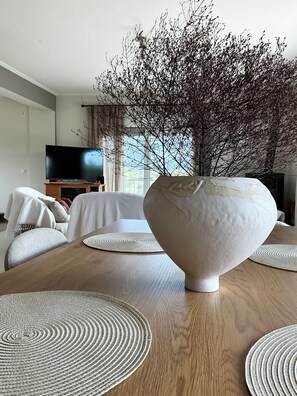 Ein modernes Wohnzimmer mit einem Holzesstisch, weißen Poufs zum Sitzen, einer großen weißen Vase mit Ästen und einem Fernseher im Hintergrund.