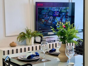 Ein Essbereich mit einem Tisch für zwei Personen, einem Fernseher, der eine Netflix-Oberfläche zeigt, und Zierpflanzen.