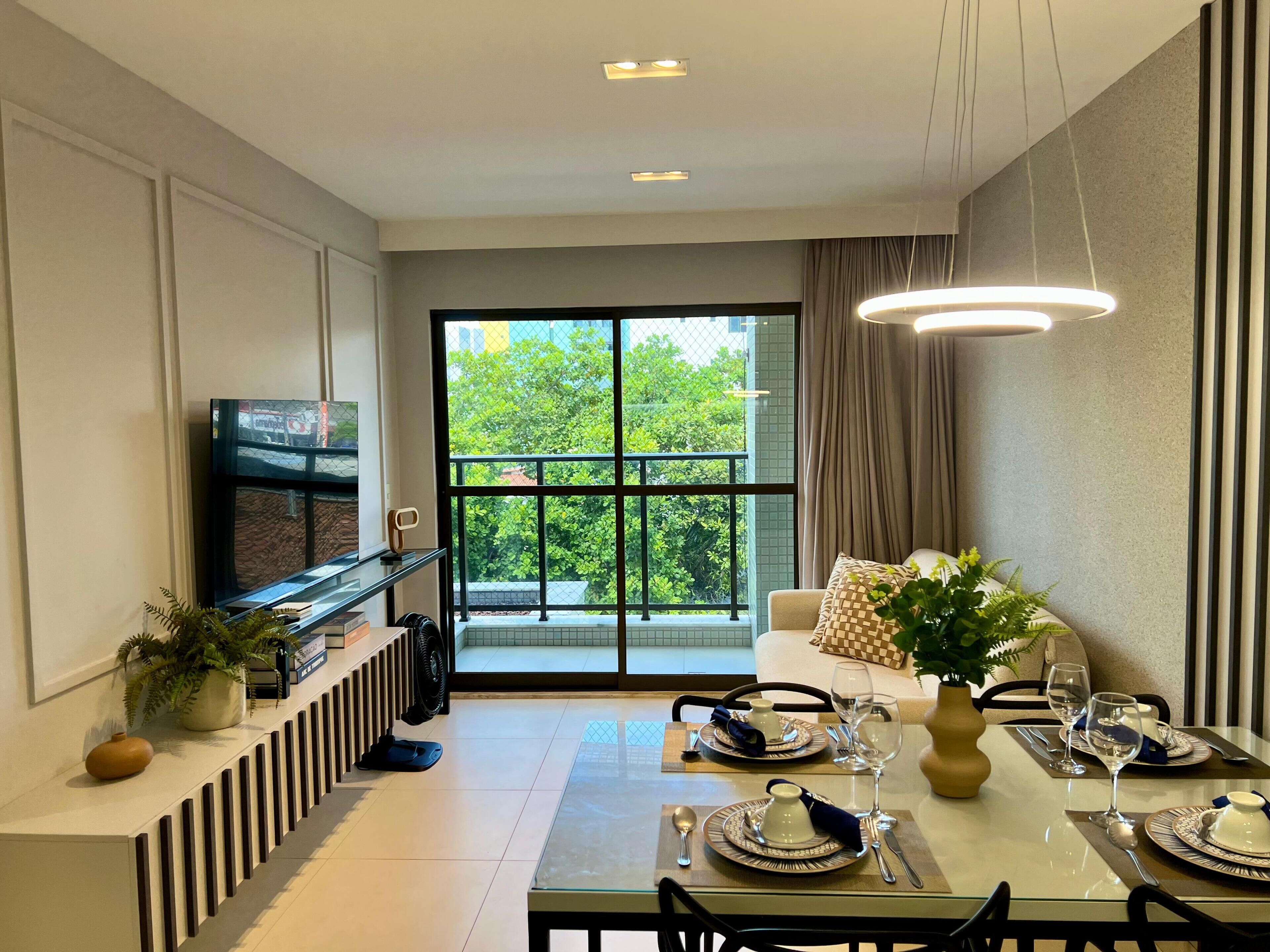 A dining area with a glass table set for two, a sofa, a TV, and a balcony with a view of trees.