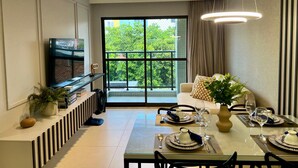 Un área de comedor con una mesa de vidrio dispuesta para dos personas, un sofá, un televisor y un balcón con vistas a árboles.