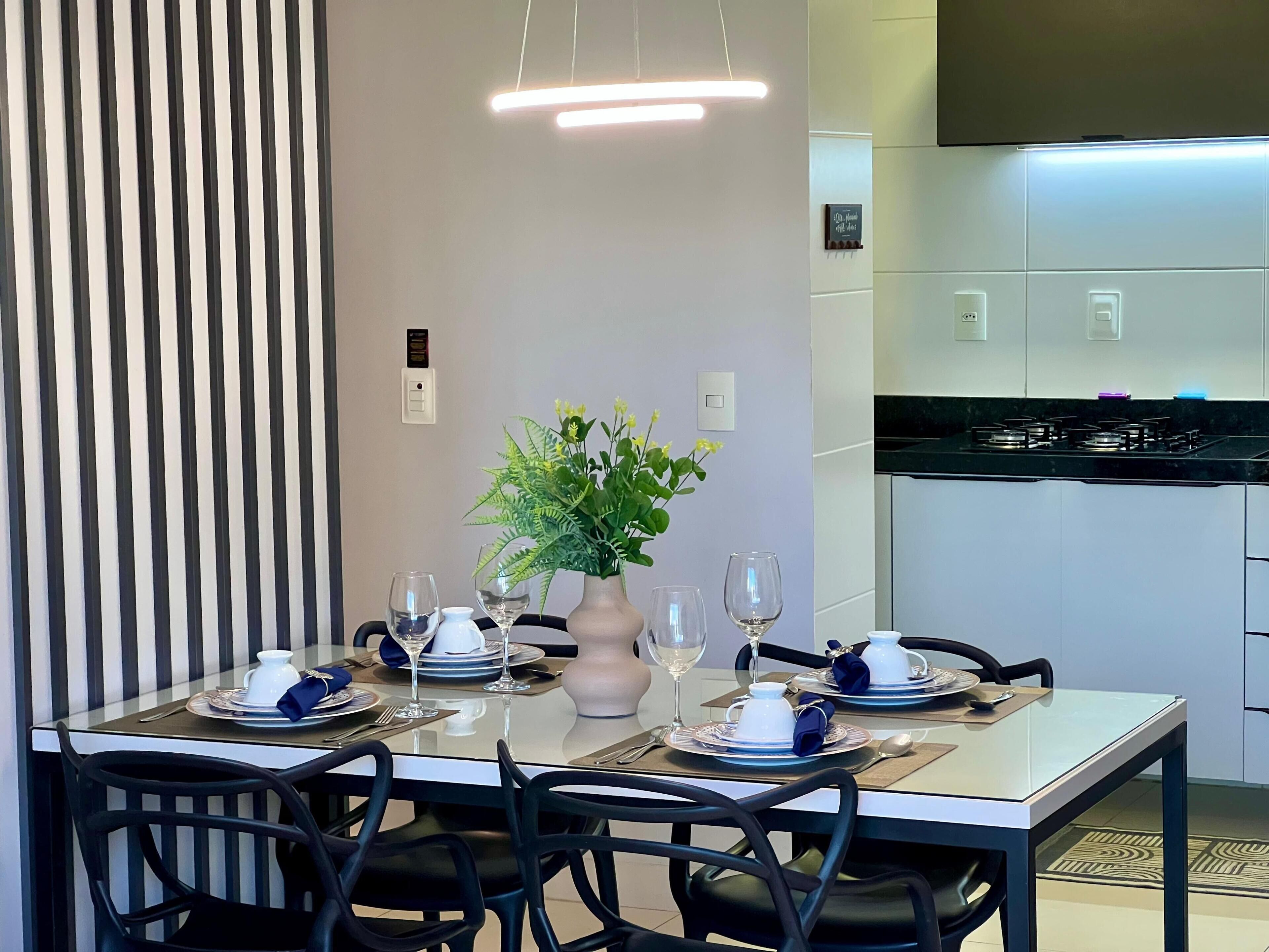 A modern dining area with a table set for two, a vase with flowers, and a kitchen in the background.