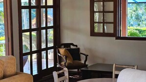 Una habitación con sofá, sillones, una mesita y una silla. Amplias ventanas con marcos de madera.