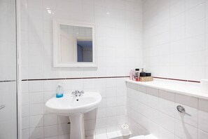 Ein Badezimmer mit weißen Fliesen, einem weißen Waschbecken, einem Spiegel und einem Ablagefach mit Pflegeprodukten.