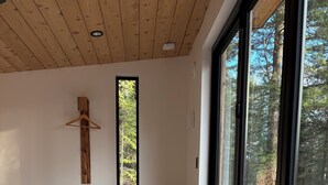 Huoneessa on puinen penkki, pieni pöytä, seinälle ripustettu puinen risti ja suuri ikkuna, josta avautuu näkymä puihin.