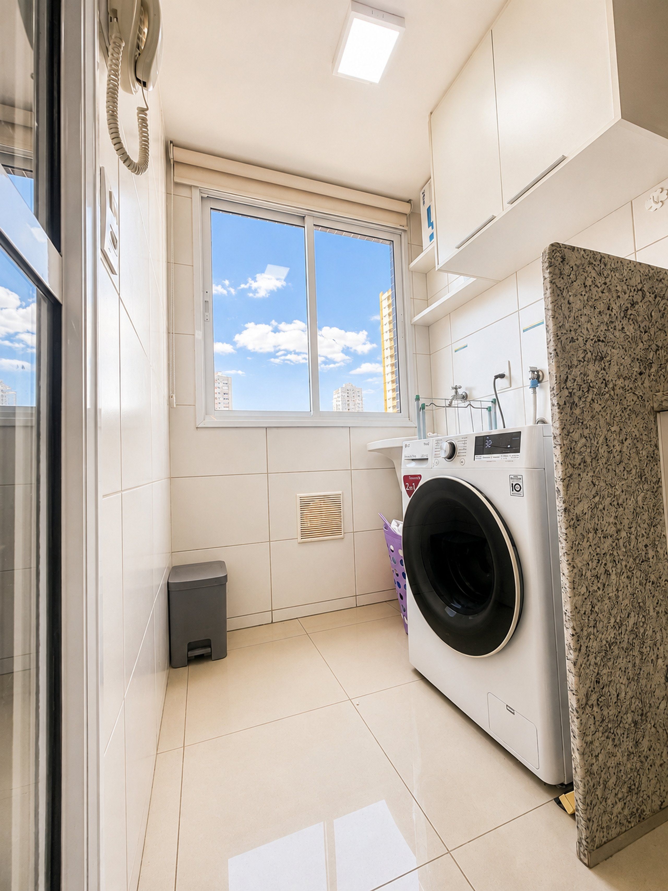 A laundry room with a washing machine, a dryer, a window with a view of buildings, and a tiled floor.