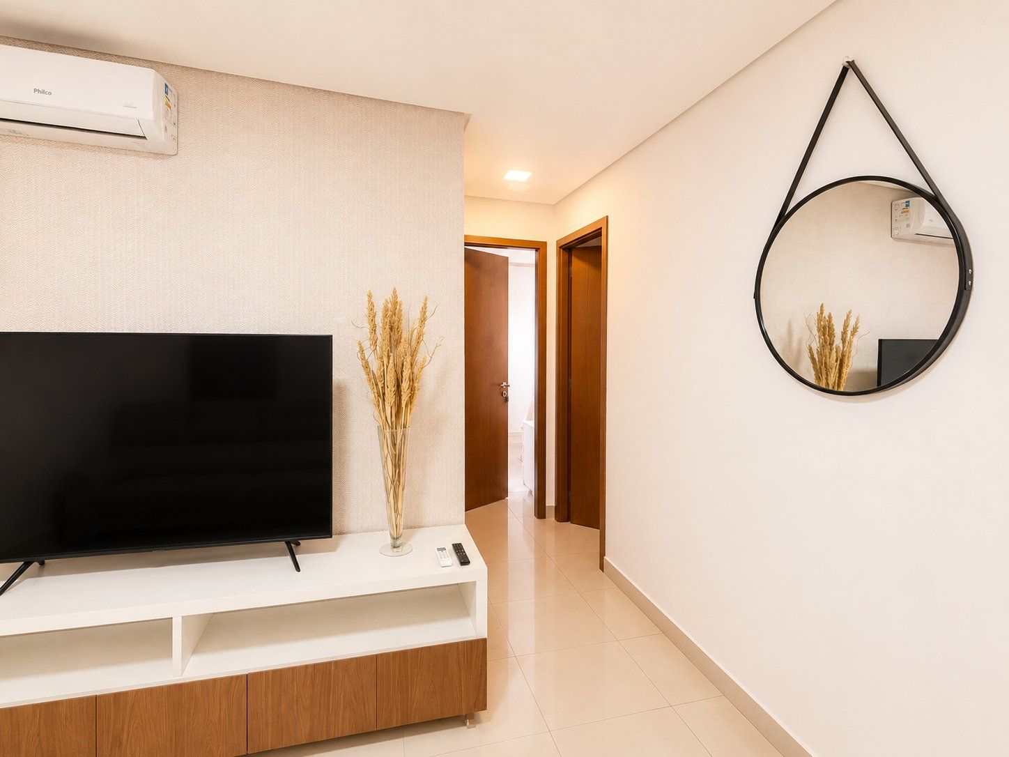 Una sala moderna con un televisor de pantalla plana sobre un mueble blanco, un aire acondicionado instalado en la pared y un espejo redondo que refleja un jarrón con plantas secas.