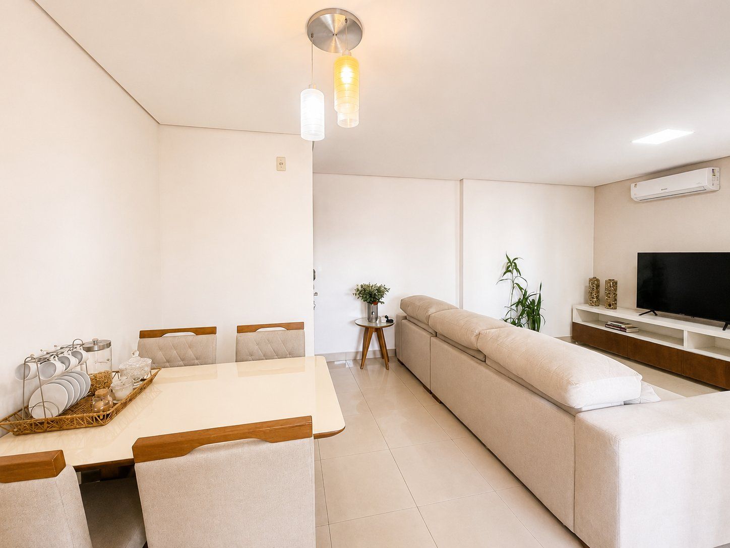 A modern living room with a sofa, a dining table with chairs, a TV on a stand, and a hanging light fixture.