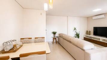 A modern living room with a sofa, a dining table with chairs, a TV on a stand, and a hanging light fixture.