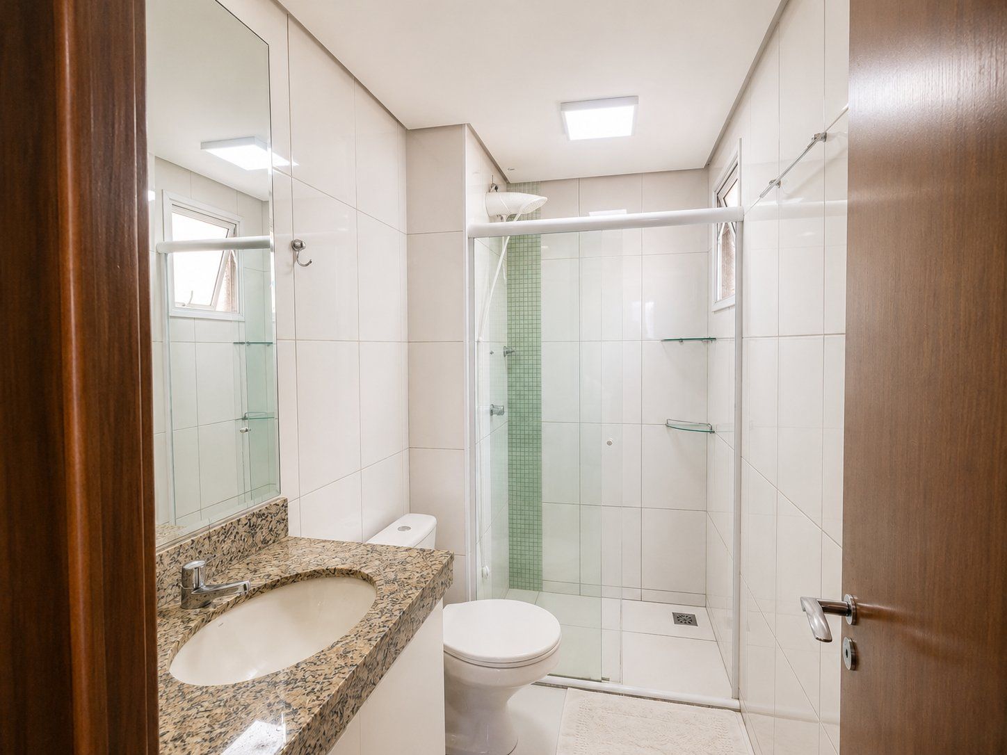 Un baño con lavamanos de encimera de granito, inodoro blanco, cabina de ducha de vidrio y puerta de madera.