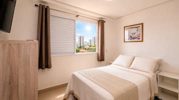 Un dormitorio con cama, televisor, ventana con cortinas, un cuadro en la pared y vistas de edificios.