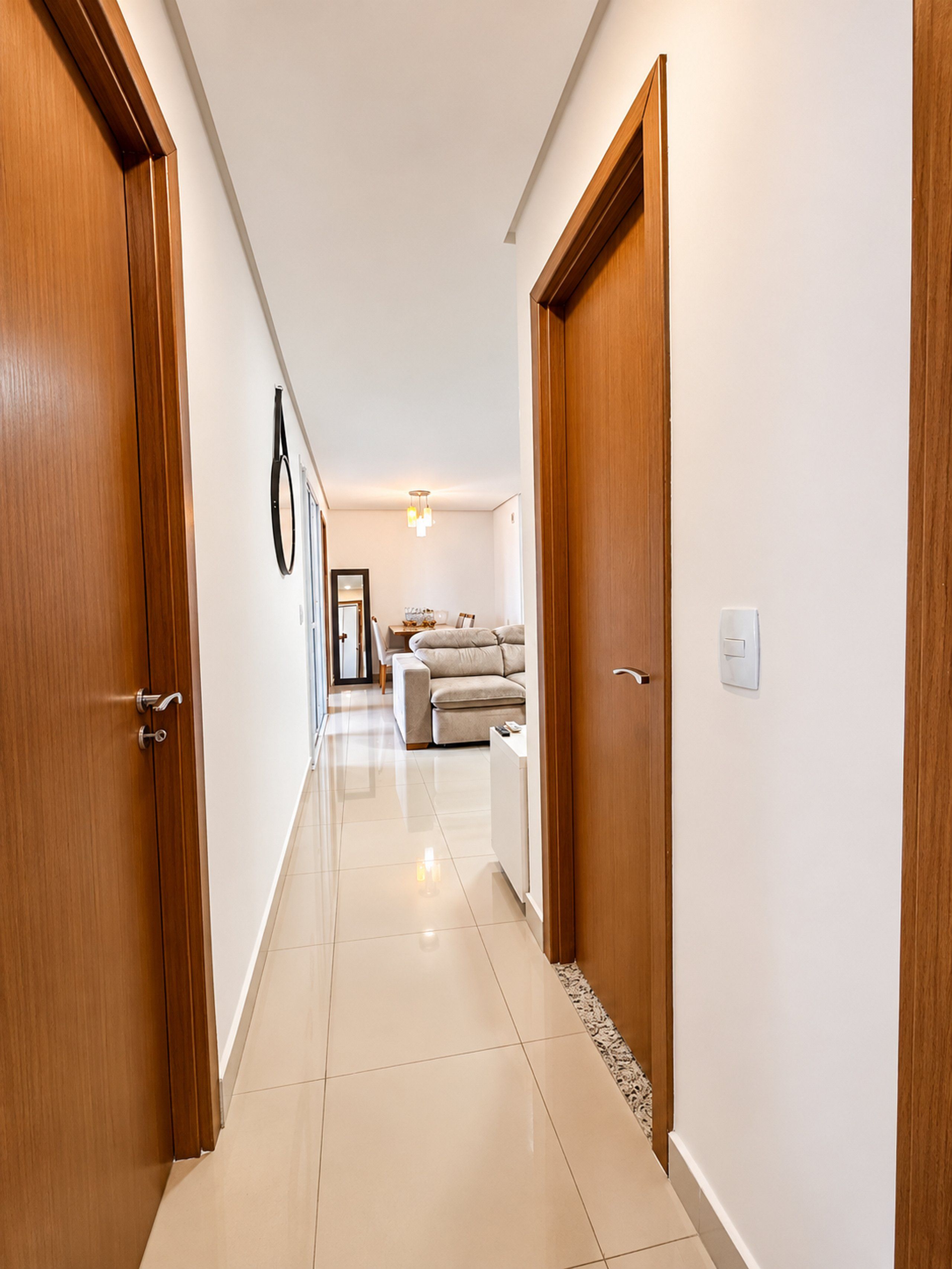 Um corredor com portas de madeira, parede branca, luminária no teto e um sofá ao longe.