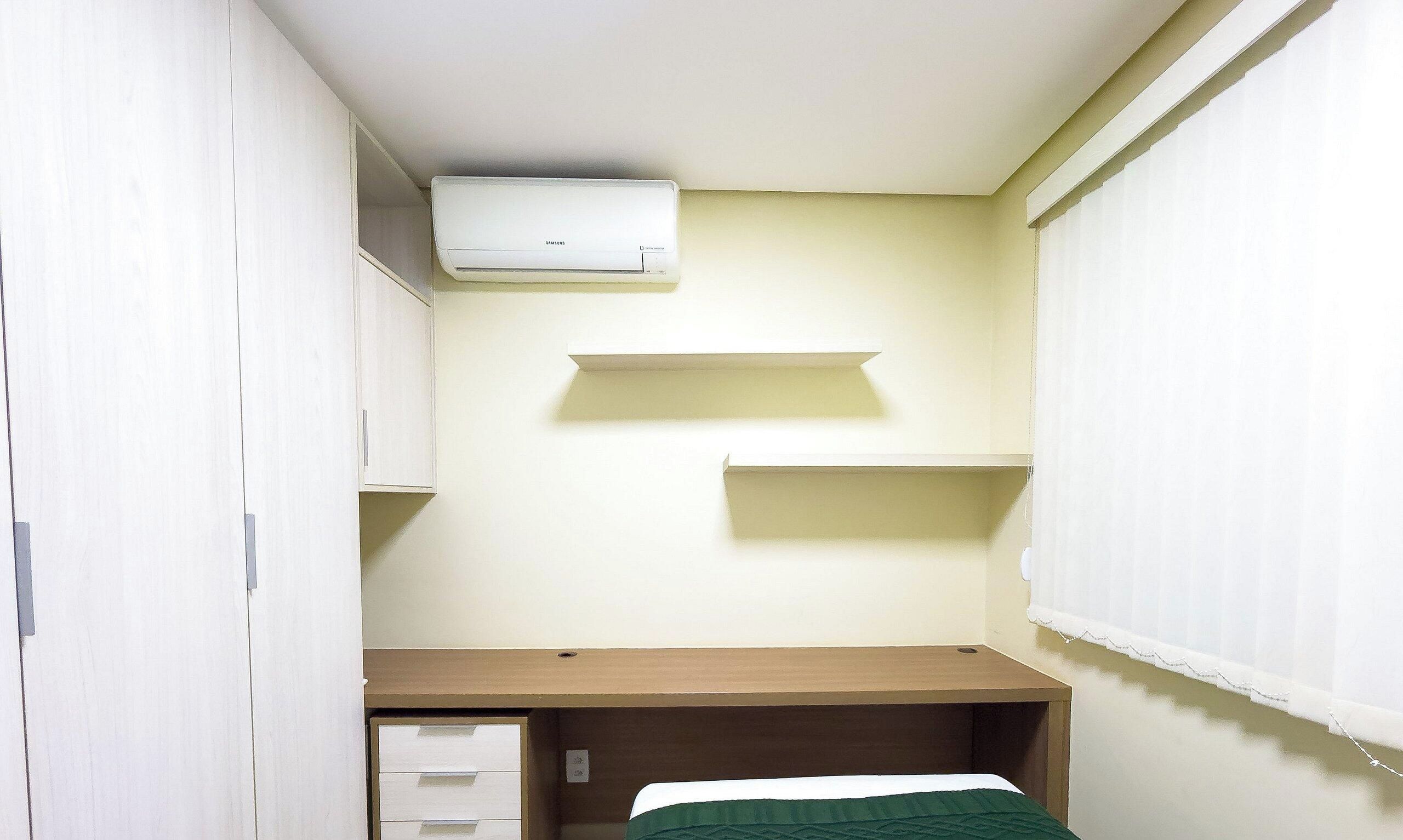 Una habitación pequeña y funcional con una cama individual, un escritorio y un equipo de aire acondicionado blanco instalado en la pared.