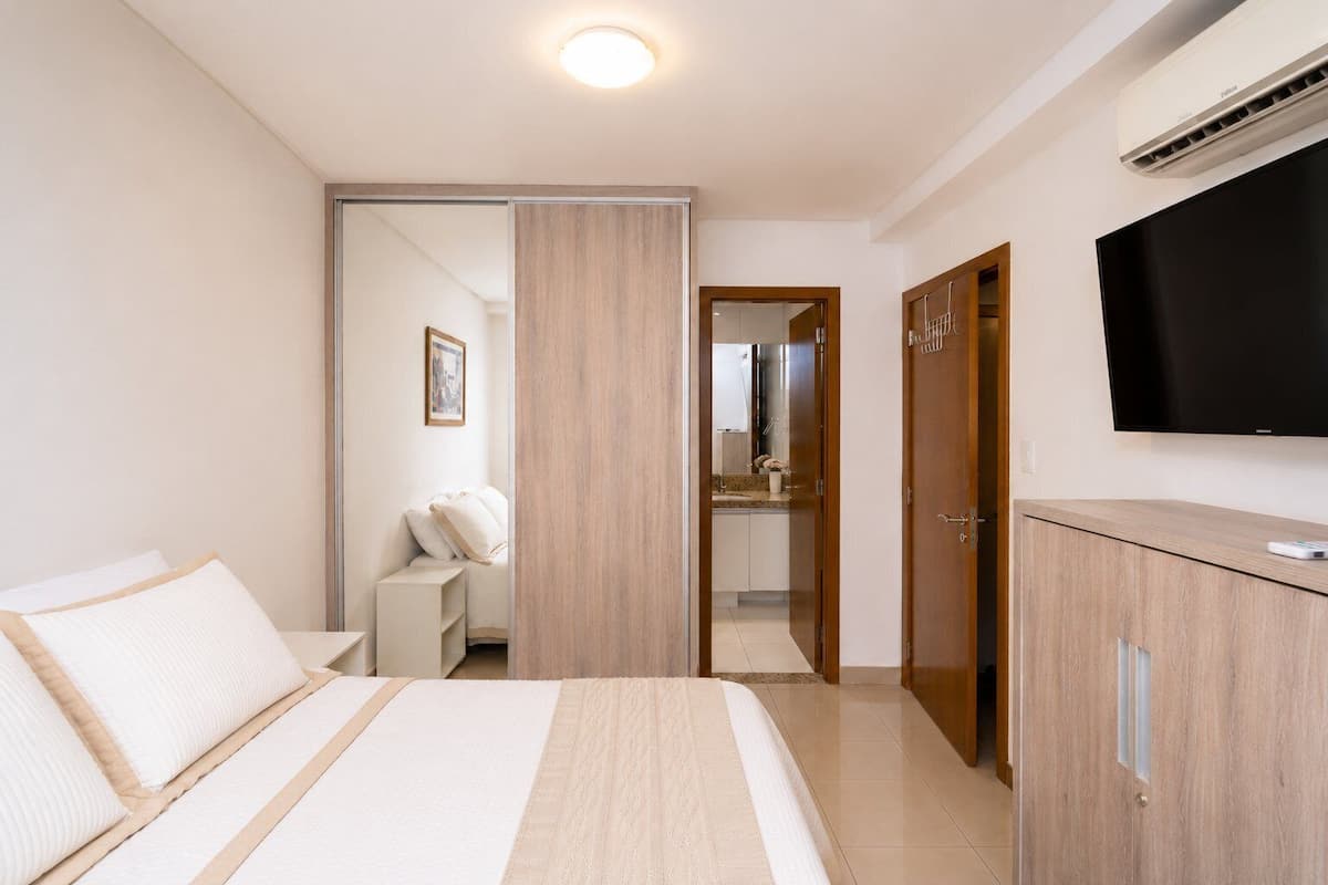 Quarto de hotel com cama, guarda-roupa, televisão e banheiro visível por uma porta aberta.