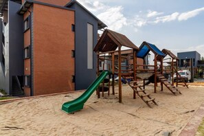 A playground with a slide and climbing structures, adjacent to a building with a brick facade. - Waterfall Hive (Midrand)