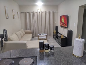A living room with a sofa, a coffee table, a TV, and a kitchen area with a stove and tea containers. - Waterfall Hive (Midrand)