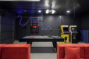 Een pooltafel met stokken, een projector en speelautomaten in een ruimte met donkere wanden en rode kussens.