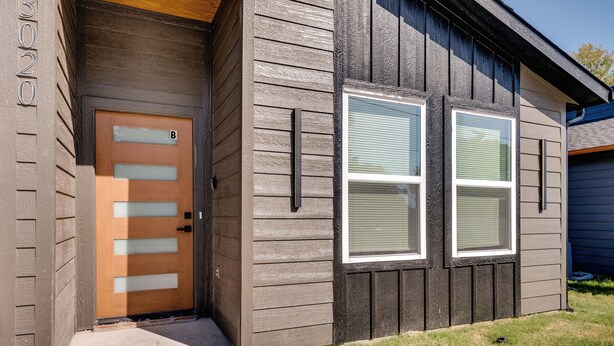 Un edificio residencial con una puerta de madera marcada 'B', dos ventanas con marcos blancos y un sendero de concreto que lleva a la entrada.