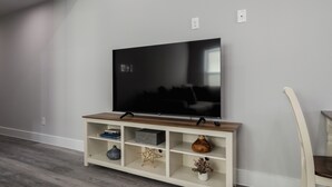 Un televisor de pantalla plana sobre un mueble de madera con estantes, una silla y una pared blanca.