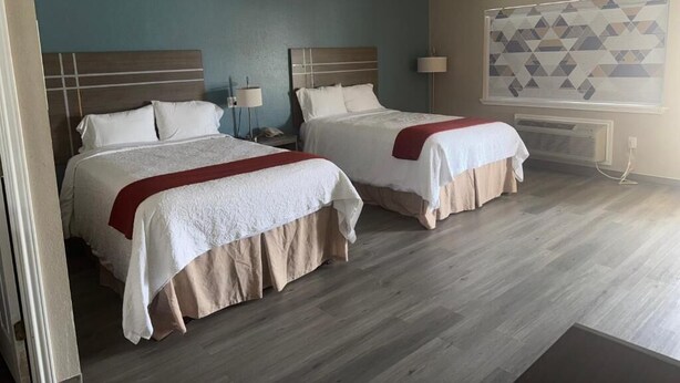 Ein Hotelzimmer mit zwei Betten, einer blauen Akzentwand, einem fensterartigen Vorhang mit geometrischem Muster und Holzboden.
