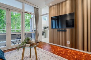 Ein modernes Wohnzimmer mit großem, an einer Holzwand montiertem Fernseher, einem Glastisch und Blick durch große Schiebetüren auf einen Außenbereich.