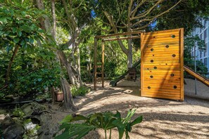 Ein Spielplatz mit Klettergerüsten und einer Rutsche, umgeben von Bäumen und einem kleinen Wasserspielplatz.