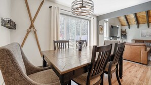 En matplats med ett träbord, stolar och en kristallkrona. I bakgrunden finns ett fönster med gardiner, en tv och en soffa.