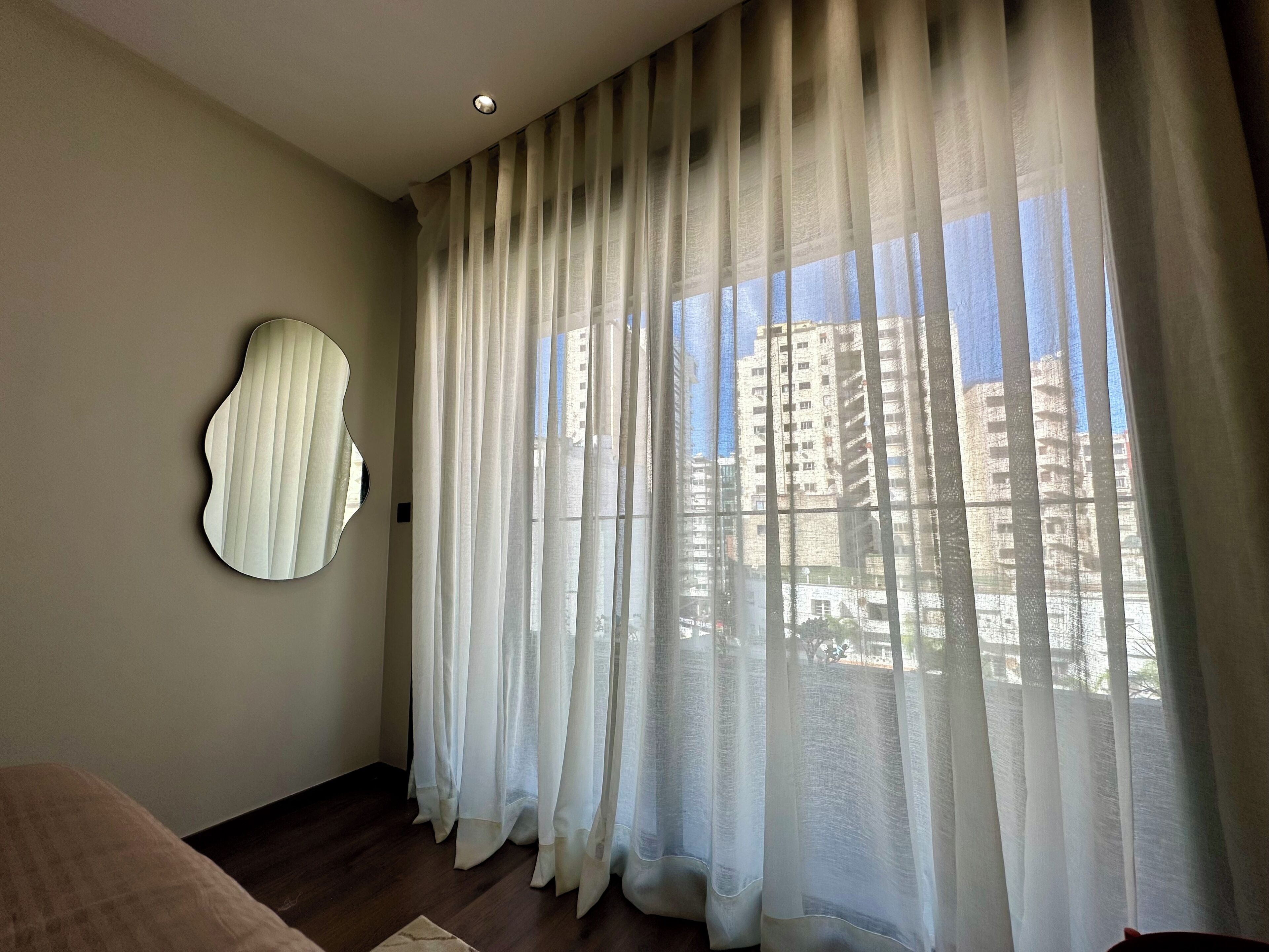 A room with sheer curtains, a round mirror on the wall, and a view of high-rise buildings.