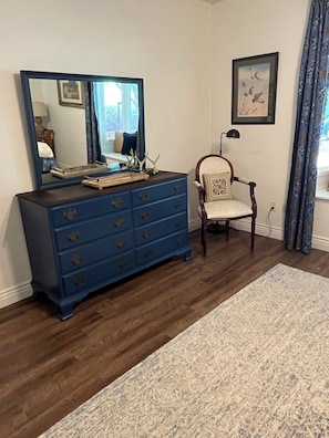 Une pièce avec une commode bleue, une chaise, un tapis, un tableau encadré et une fenêtre avec des rideaux.