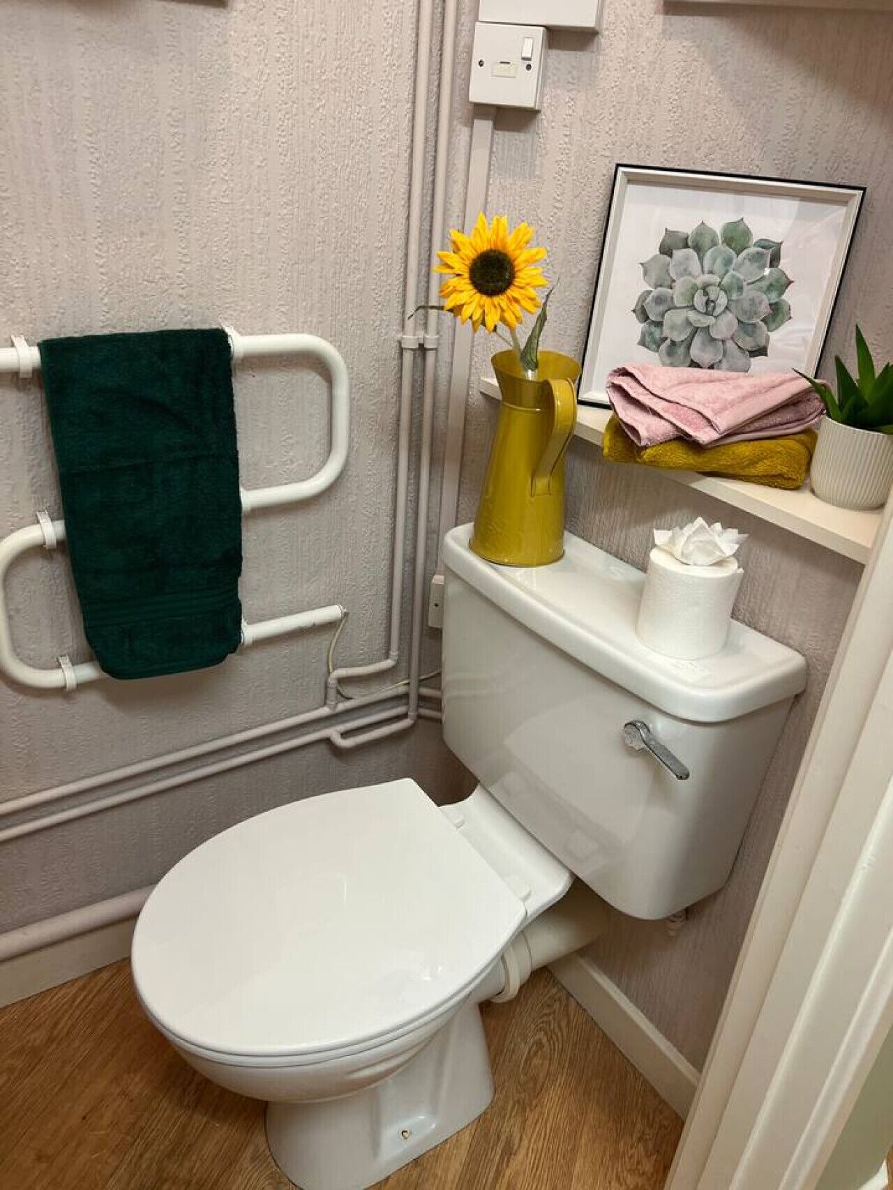 Une salle de bain avec des toilettes blanches, un porte-serviettes sur lequel est posée une serviette verte, un vase jaune contenant un tournesol et une plante succulente encadrée, placée sur une étagère.