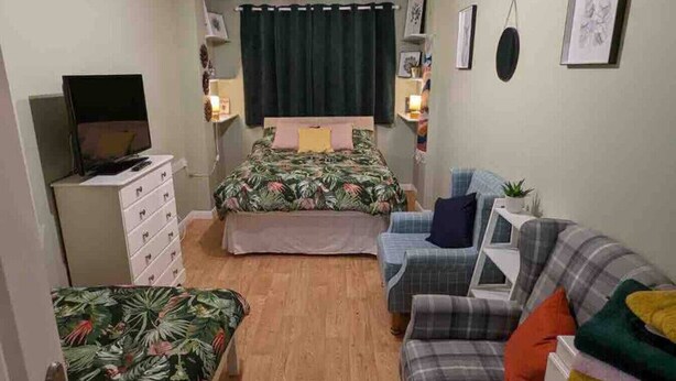 A small, furnished room with a bed, a sofa, a TV, a desk, and a chair.