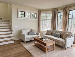 Ein Wohnzimmer mit Treppe, zwei gestreifte Sofas, einem Holztisch und großen Fenstern mit Vorhängen.
